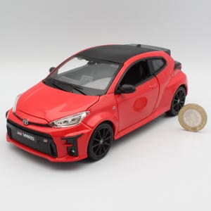 May include: A red Toyota GR Yaris scale model car featuring a black roof, black multi-spoke wheels, and a front grille with the GR Yaris logo. A circular coin is positioned next to the vehicle to provide a sense of scale for the model.