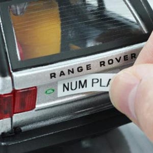 May include: A silver Range Rover toy car with black trim and red taillights. A hand is applying a white sticker that reads "NUM PLA" to the rear of the vehicle. The text "RANGE ROVER" is above the sticker.