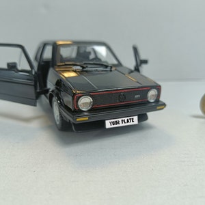 May include: A black model car with an open door, a red stripe on the front grill, and the text "YOUR PLATE" on the number plate. A coin is in the background.