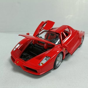 May include: A red die-cast model car with the bonnet and doors open. The car has silver wheels and a black interior. The model is a Ferrari Enzo. The car is on a white surface.