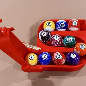 Peut inclure: Un support rouge imprimé en 3D pour boules de billard contenant onze boules de billard numérotées. Les boules sont de couleurs variées, dont le jaune, le bleu, le rouge, le vert et le violet. Le support a une poignée et une rampe.