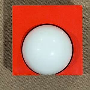 May include: A white sphere set within a bright orange square frame. The sphere is centered, creating a striking contrast against the vibrant background. The design is simple and modern, with clean lines and a minimalist aesthetic.