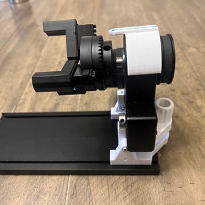 Peut inclure: Un bras robotique imprim&eacute; en 3D noir et blanc avec un syst&egrave;me d'engrenages. Le bras est mont&eacute; sur une base noire avec une plateforme blanche.