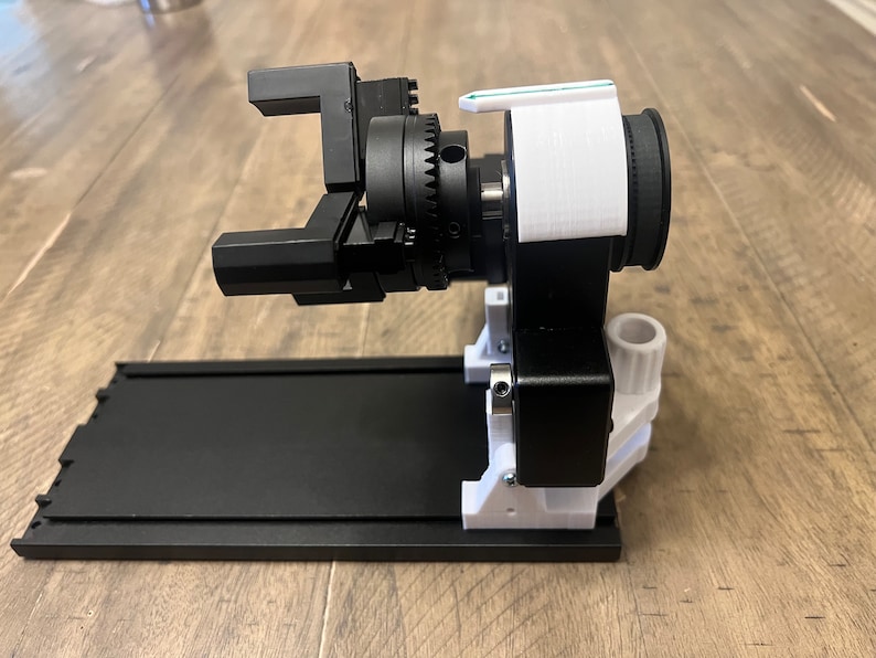 Peut inclure: Un bras robotique imprim&eacute; en 3D noir et blanc avec un syst&egrave;me d'engrenages. Le bras est mont&eacute; sur une base noire avec une plateforme blanche.