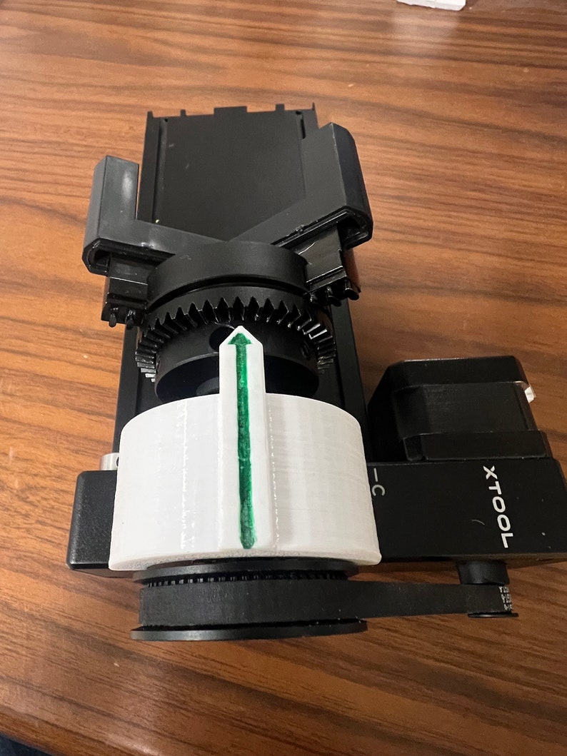 Peut inclure: Un stabilisateur de cam&eacute;ra noir et blanc avec une fl&egrave;che verte pointant vers le haut. Le stabilisateur est dot&eacute; d'un syst&egrave;me d'engrenages et d'une poign&eacute;e. La marque "TOOLX" est visible sur le c&ocirc;t&eacute;.