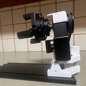 Peut inclure: Un bras robotique imprim&eacute; en 3D noir et blanc avec un syst&egrave;me d'engrenages. Le bras est mont&eacute; sur une base noire avec une pince blanche.