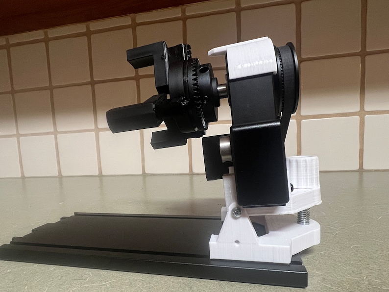 Peut inclure: Un bras robotique imprim&eacute; en 3D noir et blanc avec un syst&egrave;me d'engrenages. Le bras est mont&eacute; sur une base noire avec une pince blanche.