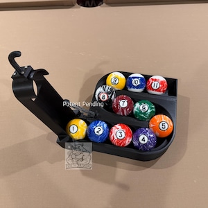 Puede incluir: Un soporte negro para bolas de billar que contiene 10 bolas numeradas. Las bolas están dispuestas en forma triangular, con los números del 1 al 11 visibles. El soporte tiene un diseño curvo y un gancho para colgar. El texto "Patent Pending" es visible.