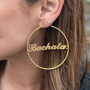 May include: Gold-colored hoop earrings with the word "Bachata" in a cursive font. The earrings are large and circular, with a heart-shaped detail at the end of the word. A small stud earring is also visible.