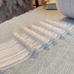 May include: A close-up of a light blue textured surface with six white, semi-transparent, cylindrical objects. The objects are arranged in a row, with a white, round decorative object in the background. A brick wall and dried flowers are also visible.