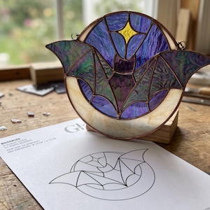 May include: A stained glass suncatcher featuring a bat silhouette centered against a crescent moon. The bat is crafted from iridescent purple glass, while the moon is made of cream-colored glass. A yellow star sits above the bat. Text on the paper reads: MOONWING, PRINT SIZE: A4 (PORTRAIT), MAX DIMENSIONS: W 19 CM x H 11 CM.