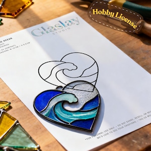 Puede incluir: Un colgante de vidrio manchado en forma de corazón con un diseño de ola en tonos azules y turquesas, sobre un papel blanco con el texto "Glaslay Studio". La imagen también muestra herramientas y piezas de vidrio, sugiriendo un proyecto artesanal.