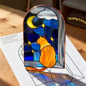 May include: Stained glass window art featuring an orange cat gazing at a moon and stars. The window is framed in copper and hangs from a chain. The design includes blue, yellow, and gray glass. The text "THE NIGHT WATCHER" is visible.