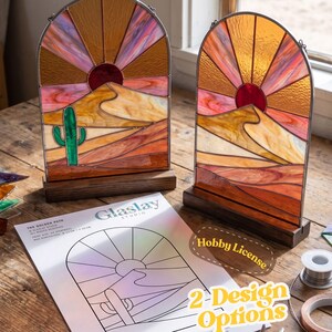 May include: Two arched stained glass panels featuring a desert landscape with a sun, sand dunes, and a green cactus, each set in a dark wooden base. A printed paper pattern labelled The Golden Path and Glaslay Studio is visible in the foreground.