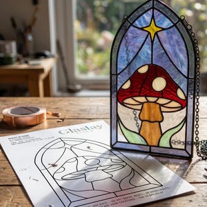 May include: A stained glass panel featuring a red mushroom with white spots under a yellow star, set in a gothic arch frame. Beside it lies a paper pattern sheet from Glaslay Studio titled Night Bloom with assembly instructions.