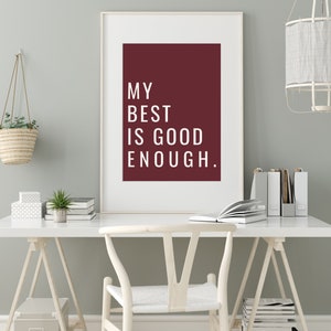 Może przedstawiać: Biały obraz w ramie z bordowym tłem i białym tekstem, kt&oacute;ry brzmi "My best is good enough." Obraz wisi na jasnoszarej ścianie nad białym biurkiem z białym krzesłem. Na biurku znajduje się wisząca doniczka z plecionym koszem i mała doniczkowa roślina.