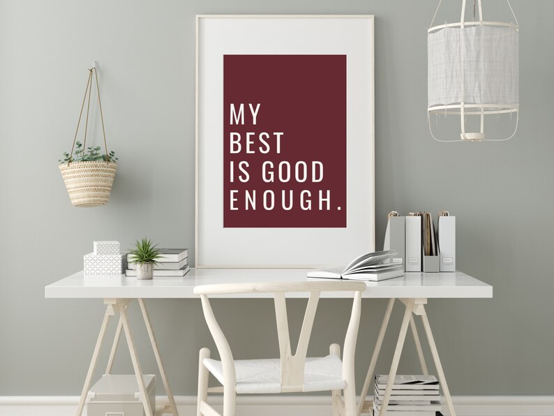 Może przedstawiać: Biały obraz w ramie z bordowym tłem i białym tekstem, kt&oacute;ry brzmi "My best is good enough." Obraz wisi na jasnoszarej ścianie nad białym biurkiem z białym krzesłem. Na biurku znajduje się wisząca doniczka z plecionym koszem i mała doniczkowa roślina.