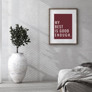 Może przedstawiać: Wydruk w ramie z bordowym tłem i białym tekstem, kt&oacute;ry brzmi "My best is good enough." Wydruk wisi na białej ścianie z rośliną w białej doniczce i szarym ł&oacute;żkiem na pierwszym planie.