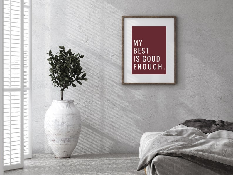 Może przedstawiać: Wydruk w ramie z bordowym tłem i białym tekstem, kt&oacute;ry brzmi "My best is good enough." Wydruk wisi na białej ścianie z rośliną w białej doniczce i szarym ł&oacute;żkiem na pierwszym planie.