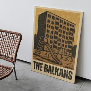 May include: A brown woven chair with metal legs sits next to a framed print. The print features a black and tan illustration of a building with a playground and the text "THE BALKANS".