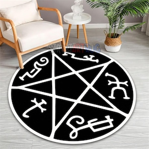 May include: A round black rug featuring a white pentagram design with stylized symbols. The rug is placed on a light-colored wooden floor, with a white chair and side table in the background. A potted plant is also visible.