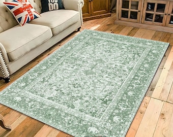 May include: A rectangular, sage green area rug with an ornate, floral pattern. The rug has a white border and is placed on a hardwood floor. The rug is in a living room setting with a sofa and wooden furniture.