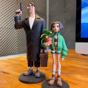 Può includere: Due figurine stilizzate, una maschile con una pistola e l'altra femminile con una pianta in vaso. La figura maschile indossa un cappotto nero e la femminile una giacca verde. Entrambe sono su basi circolari nere.