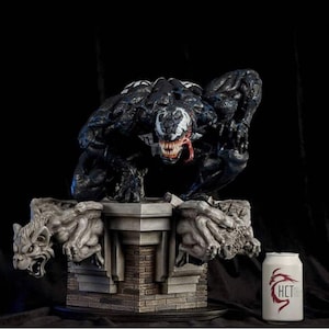 May include: A detailed statue of the Marvel Comics character Venom, in a crouching pose, atop a stone base with gargoyle figures. The statue is predominantly black with white accents and a red tongue. A white can with red text is to the right.