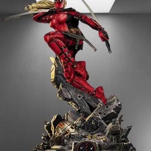 May include: A dynamic statue of a character in a red and black suit, poised in a combat stance with two swords. The figure has a mask and blonde hair. The statue is mounted on a detailed, rocky base with metallic accents, showcasing a superhero theme.