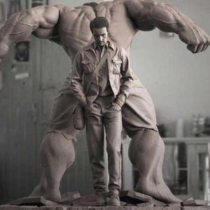 May include: A monochrome image showing a man standing before a large, muscular sculpture. The man wears a jacket and trousers. The sculpture is highly detailed, with a textured surface. The setting appears to be an artist's studio.