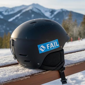 May include: Black ski helmet with a sticker that says "FAIL" in white text on a blue background. The helmet is sitting on a snow-covered wooden bench with a mountain range in the background.