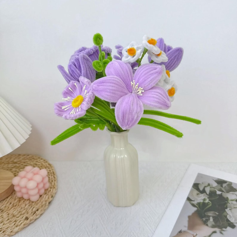 以下が含まれることがあります： 白い花瓶に入った手作りの花のブーケ。花は紫、白、緑の色合いの柔らかくベルベットのような素材で作られています。アレンジメントには、大きな星型の花と、小さくデイジーのような花が含まれています。