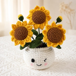Könnte beinhalten: Ein gehäkeltes Sonnenblumenarrangement mit drei großen gelben Blüten mit braunen Mitten und zwei kleinen grünen Knospen. Die Blumen stecken in einem runden, weißen Häkeltopf mit einem gestickten Gesicht mit schwarzen Augen, Lächeln und rosa Wangen.
