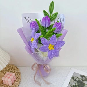 以下が含まれることがあります： 紫色のベルベットの花束。チューリップや星型の花で、黄色の中心があります。ラベンダー色の紙で包まれ、リボンで結ばれています。緑色のベルベットの葉と、紙の裏には「You're LOVE」の文字があります。