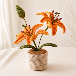 May include: A crocheted orange lily plant with two open flowers and one green bud. The plant has dark green stems and leaves, with dark red-tipped stamens. It is potted in a textured, beige crocheted pot with a brown circular top surface.