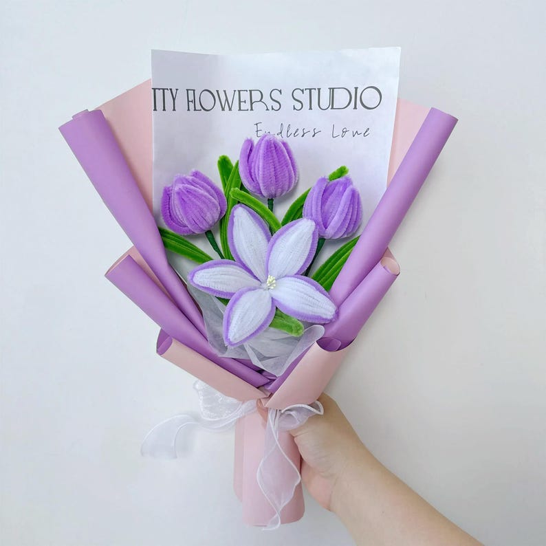 以下が含まれることがあります： 手作りの花のブーケ。紫色のチューリップと白と紫の花が特徴で、ピンクとラベンダー色の紙で包まれています。ブーケは白いリボンで結ばれ、「PRETTY FLOWERS STUDIO Endless Love」と書かれた紙が含まれています。