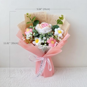 Puede incluir: Un ramo de flores de ganchillo, envuelto en papel rosa y atado con una cinta. El ramo incluye flores blancas, rosas y amarillas, y mide aproximadamente 30 cm de alto y 25 cm de ancho. La cinta dice "JUST FOR YOU".
