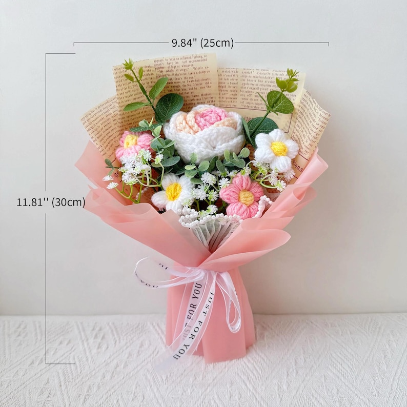 Puede incluir: Un ramo de flores de ganchillo, envuelto en papel rosa y atado con una cinta. El ramo incluye flores blancas, rosas y amarillas, y mide aproximadamente 30 cm de alto y 25 cm de ancho. La cinta dice "JUST FOR YOU".