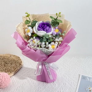 Puede incluir: Un ramo de flores de ganchillo hechas a mano en tonos morados, blancos y amarillos, envuelto en papel rosa y atado con una cinta que dice "Just for You". El ramo incluye una gran rosa morada y varias flores peque&ntilde;as en forma de margarita.