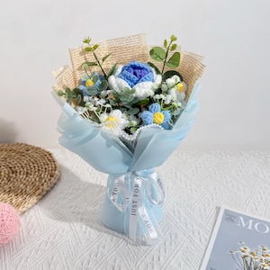 Puede incluir: Un ramo de flores de ganchillo hechas a mano en tonos azules, blancos y amarillos, envuelto en papel azul claro y atado con una cinta que dice "Just for You". El ramo incluye una gran rosa azul y blanca y otras flores m&aacute;s peque&ntilde;as.