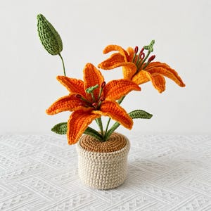 Peut inclure: Une plante lys orange crochetée avec deux fleurs ouvertes et un bouton fermé, dotée de tiges et de feuilles vertes. La plante est placée dans un pot crocheté beige avec un dessus marron, mettant en valeur des textures de fil détaillées.