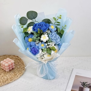 Puede incluir: Un ramo de flores de ganchillo hechas a mano en tonos azules, blancos y amarillos, envuelto en papel azul claro y atado con una cinta blanca. El ramo incluye rosas, margaritas y otros elementos florales, con acentos de follaje verde.