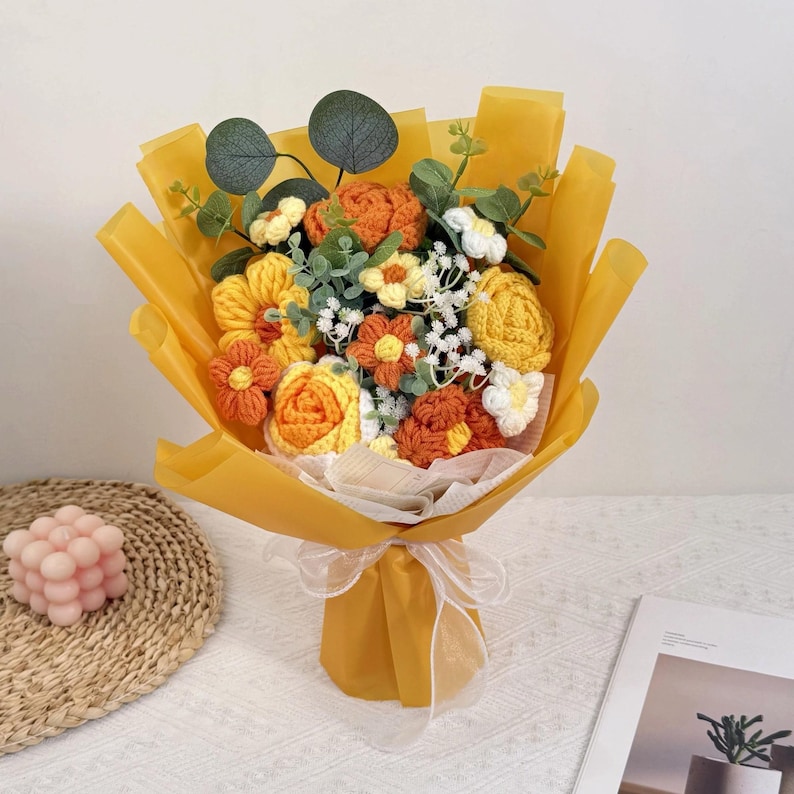 Puede incluir: Un ramo de flores de ganchillo en tonos amarillos, naranjas y blancos, envuelto en papel amarillo brillante y atado con una cinta blanca. El ramo incluye varios tipos de flores y follaje verde.