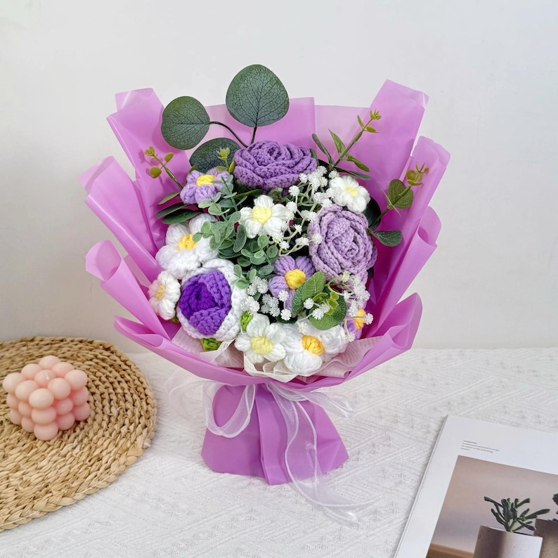 Puede incluir: Un ramo artesanal de flores de ganchillo en tonos morados, blancos y verdes, envuelto en papel lavanda. El ramo presenta rosas, margaritas y otros elementos florales, atado con una cinta blanca.