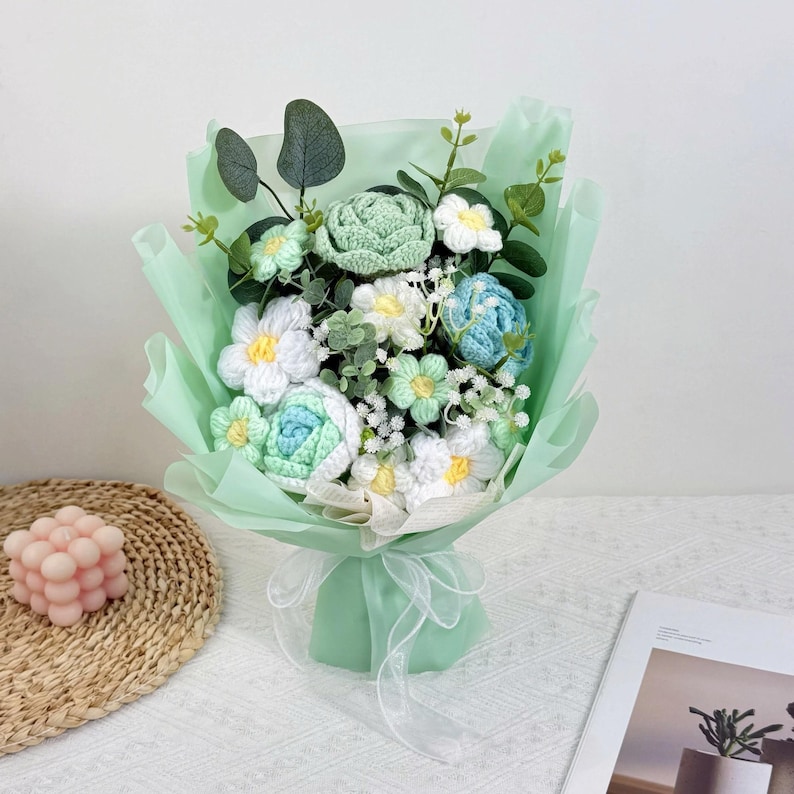 Puede incluir: Un ramo de flores de ganchillo hechas a mano en tonos blancos, verdes y azules, envuelto en papel verde claro y atado con una cinta blanca. El ramo incluye varios tipos de flores y vegetaci&oacute;n, creando un arreglo delicado y texturizado.