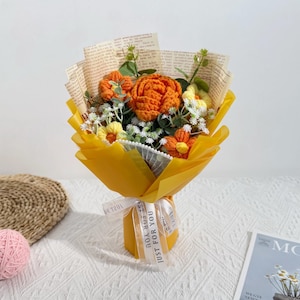 Puede incluir: Un ramo de flores de ganchillo en tonos naranja, amarillo y blanco, envuelto en papel amarillo y peri&oacute;dico vintage. El ramo est&aacute; atado con una cinta blanca que dice "JUST FOR YOU".