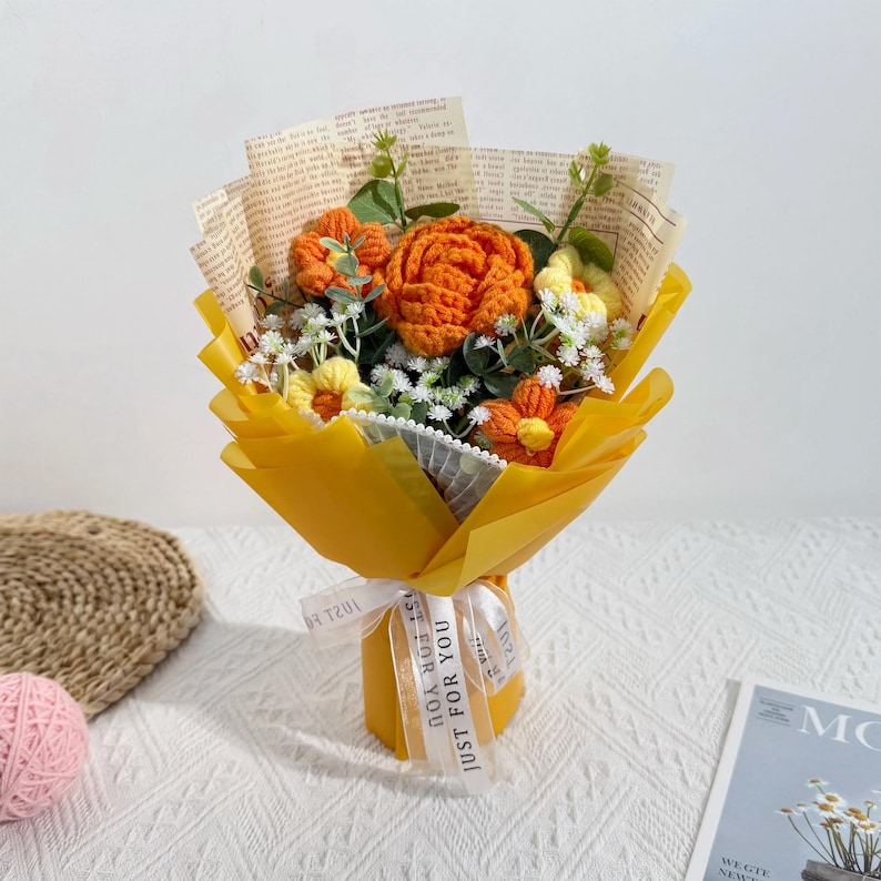Puede incluir: Un ramo de flores de ganchillo en tonos naranja, amarillo y blanco, envuelto en papel amarillo y peri&oacute;dico vintage. El ramo est&aacute; atado con una cinta blanca que dice "JUST FOR YOU".