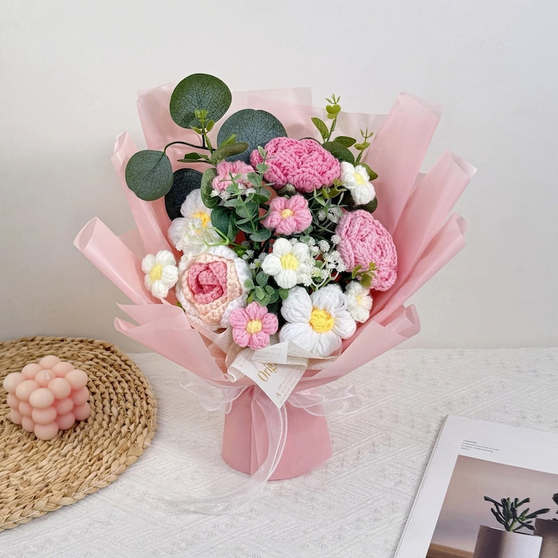 Puede incluir: Un ramo de flores de ganchillo hechas a mano en tonos rosa, blanco y verde, envuelto en papel rosa. El ramo incluye rosas, margaritas y otros elementos florales, atados con una cinta blanca. Una vela decorativa y una revista est&aacute;n en el fondo.