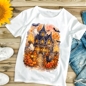 May include: A white t-shirt featuring a colorful graphic print of a haunted house at night. The illustration includes a black cat, two white ghosts, bats, a full moon, orange pumpkins, and autumn foliage surrounding the house and stone path.