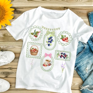 May include: A white short-sleeved t-shirt featuring a graphic print of various framed illustrations including a basket of strawberries, blueberries, a teacup, and assorted berries, accented with a small green and white checkered bunting banner.